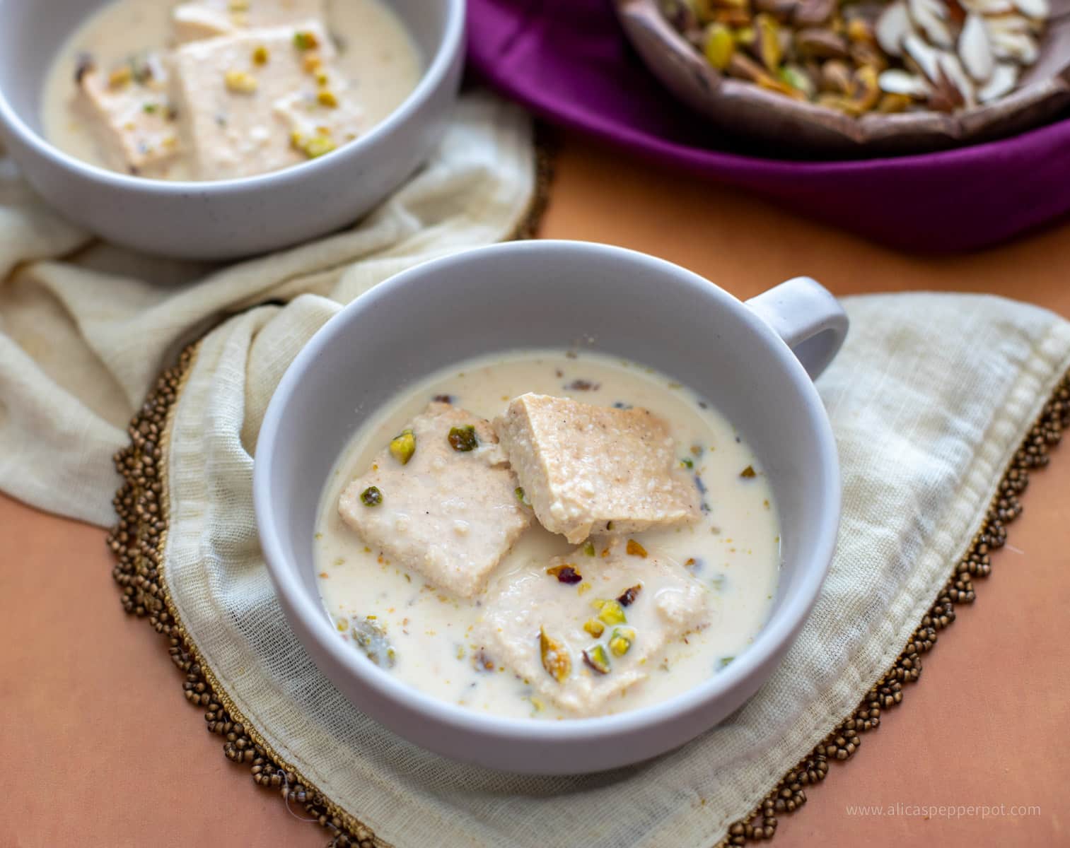 Rasmalai in two grey bowls topped with pistachios sitting on an ivory napkin with a wooden bowl of almonds and pistachios behind.