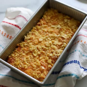 macaroni pie after coming out of oven golden brown topped with crackers in a loaf pan on a towel that has red orange blue and teal stripes