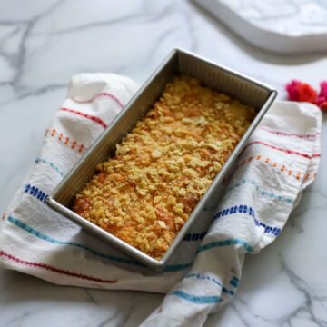macaroni pie after coming out of oven golden brown topped with crackers in a loaf pan on a towel that has red orange blue and teal stripes