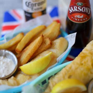 This is an image of British fish and chips arranged in a mesh basket topped with a lemon and served alongside tartar sauce and malt vinegar