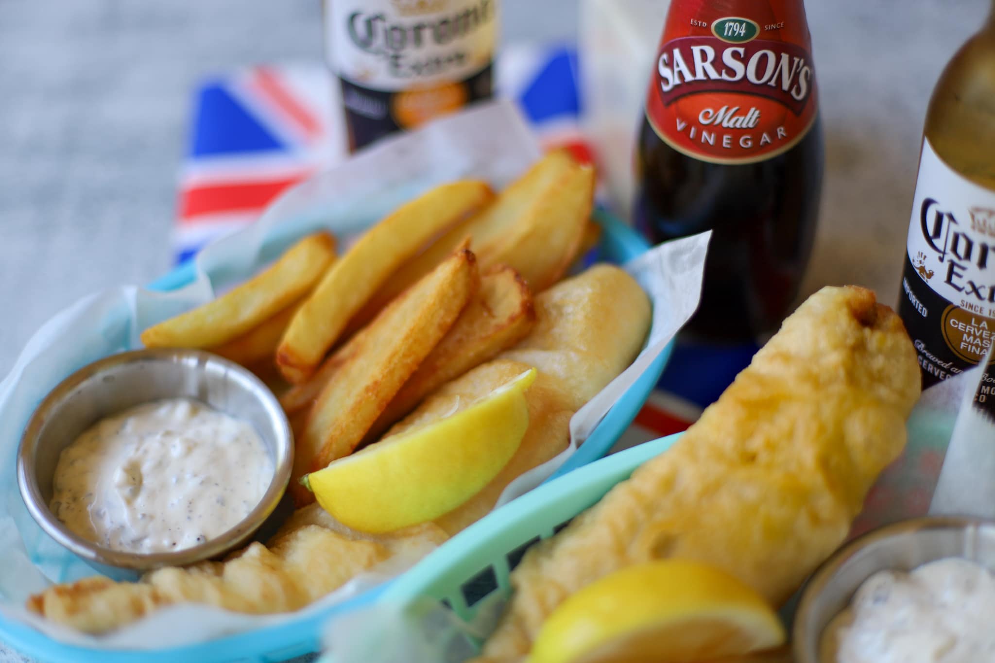 This is an image of British fish and chips arranged in a mesh basket topped with a lemon and served alongside tartar sauce and malt vinegar