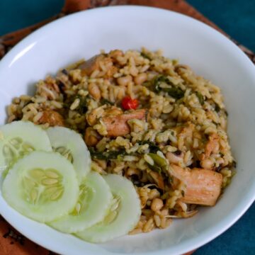 This photo shows guyanese cookup rice in a white plate with chicken in it and spinach it also has sliced cucumbers on the side